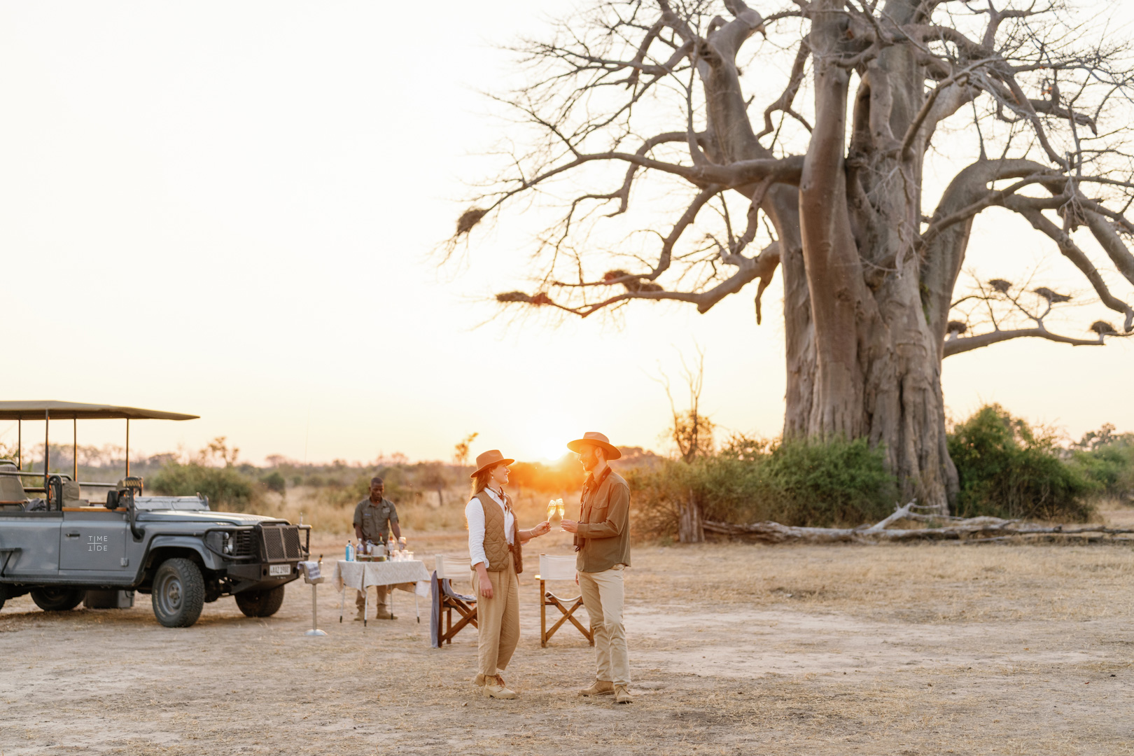 A couple clinking their glasses on a romantic sundowner game drive stop next to a magnificent baobab tree with Time + Tide Safaris in the South Luangwa Zambia