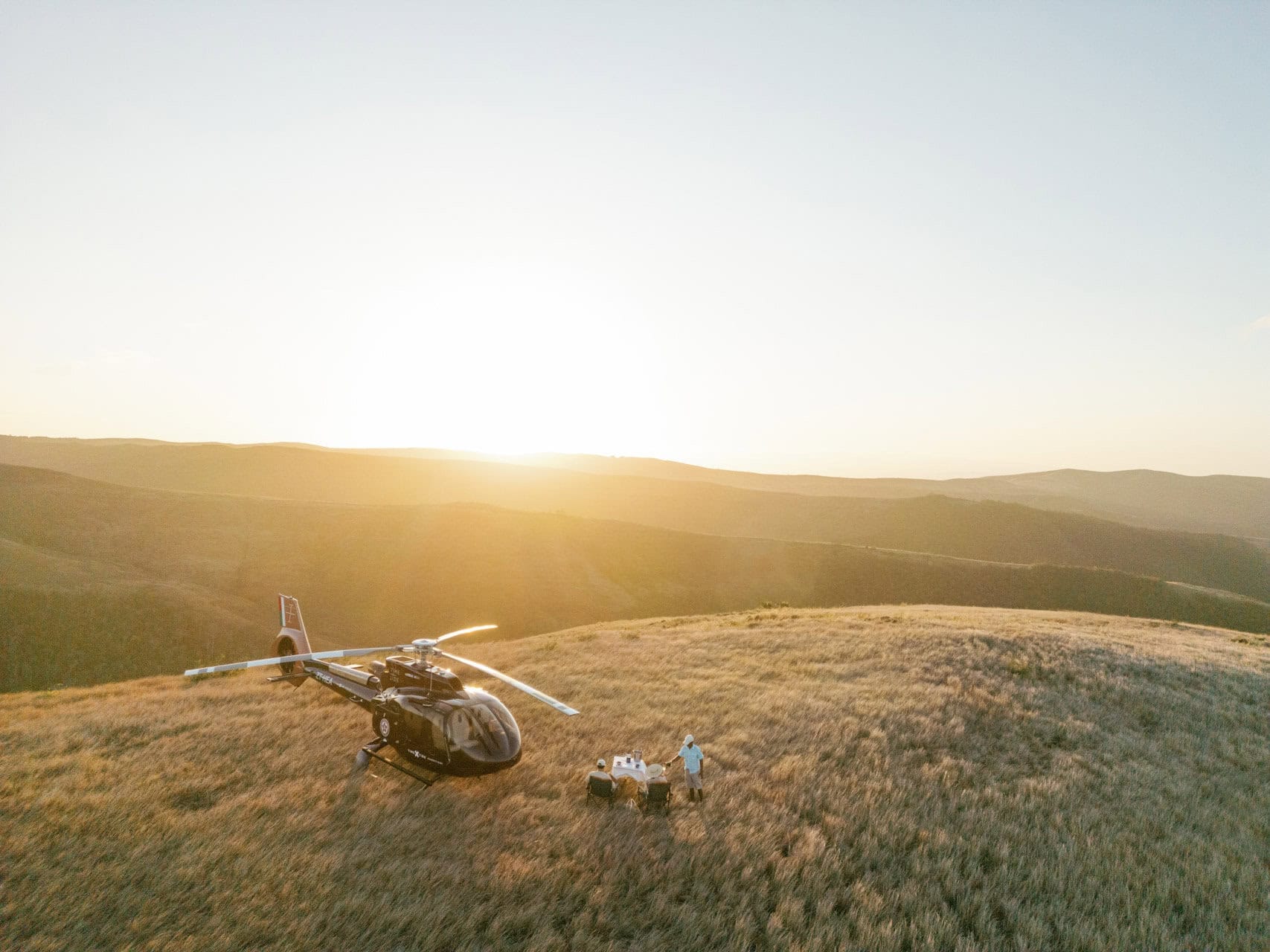 Two guests enjoying their sundowner setup on a ridge and next to the parked helicopter at sunset with Miavana by Time + Tide Madagascar