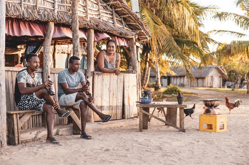 Two men sitting outside on a bench and a women standing inside the bar at Pama Salance at Miavana by Time + Tide Madagascar
