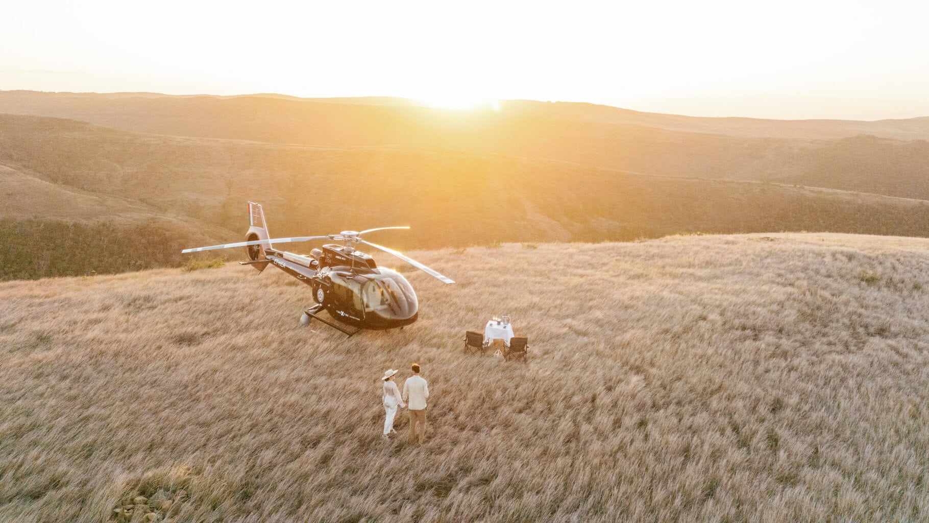 Two guests walking towards their sundowner setup on a ridge and next to the parked helicopter at sunset with Miavana, Madagascar